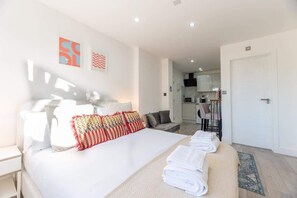 A modern hotel room with a large bed, a sitting area, a kitchenette, and a bathroom.