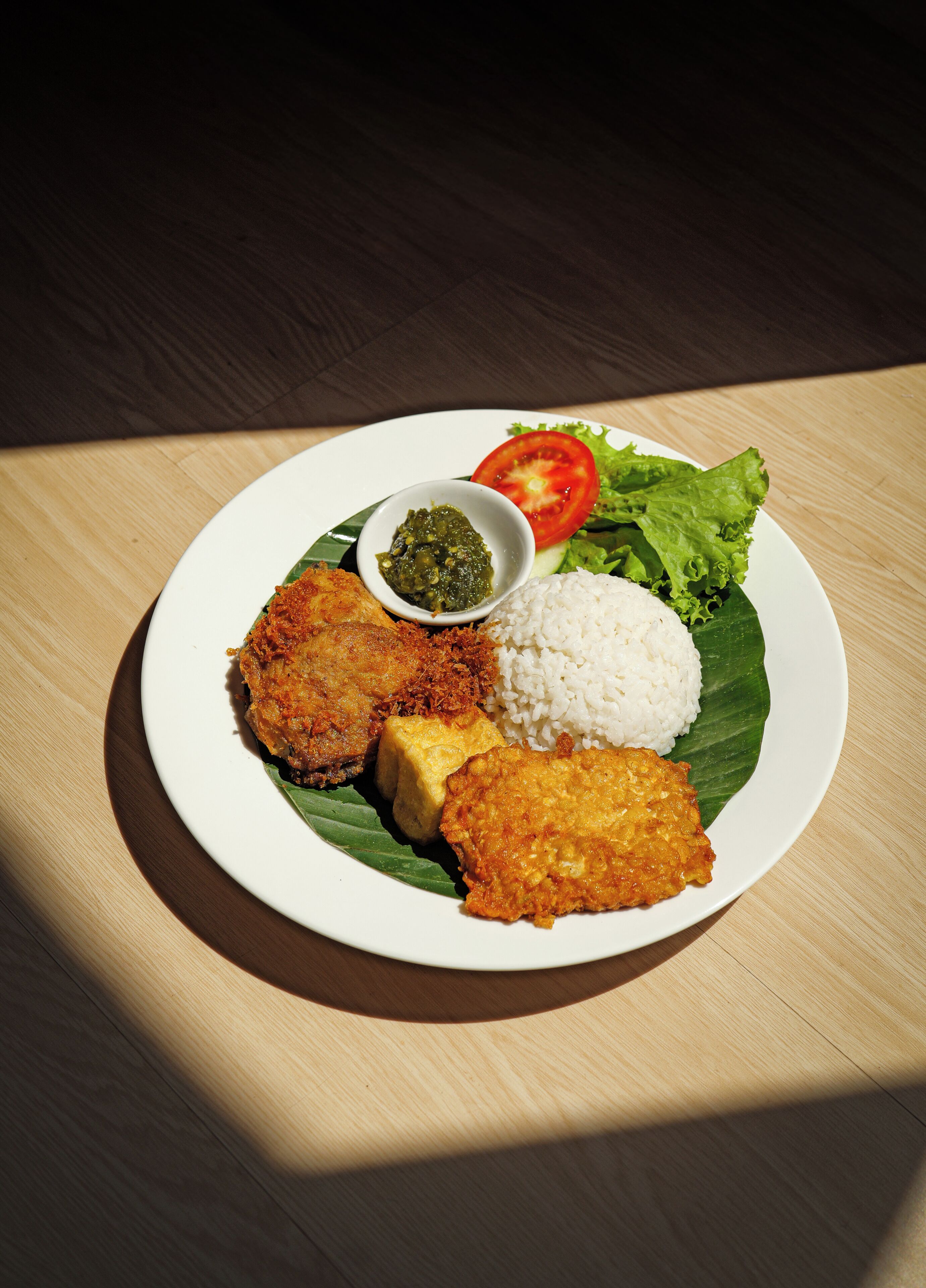 Prato de comida com arroz, um alimento frito, um pedaço de tofu e uma pequena tigela de molho, servido em um prato branco com uma folha.