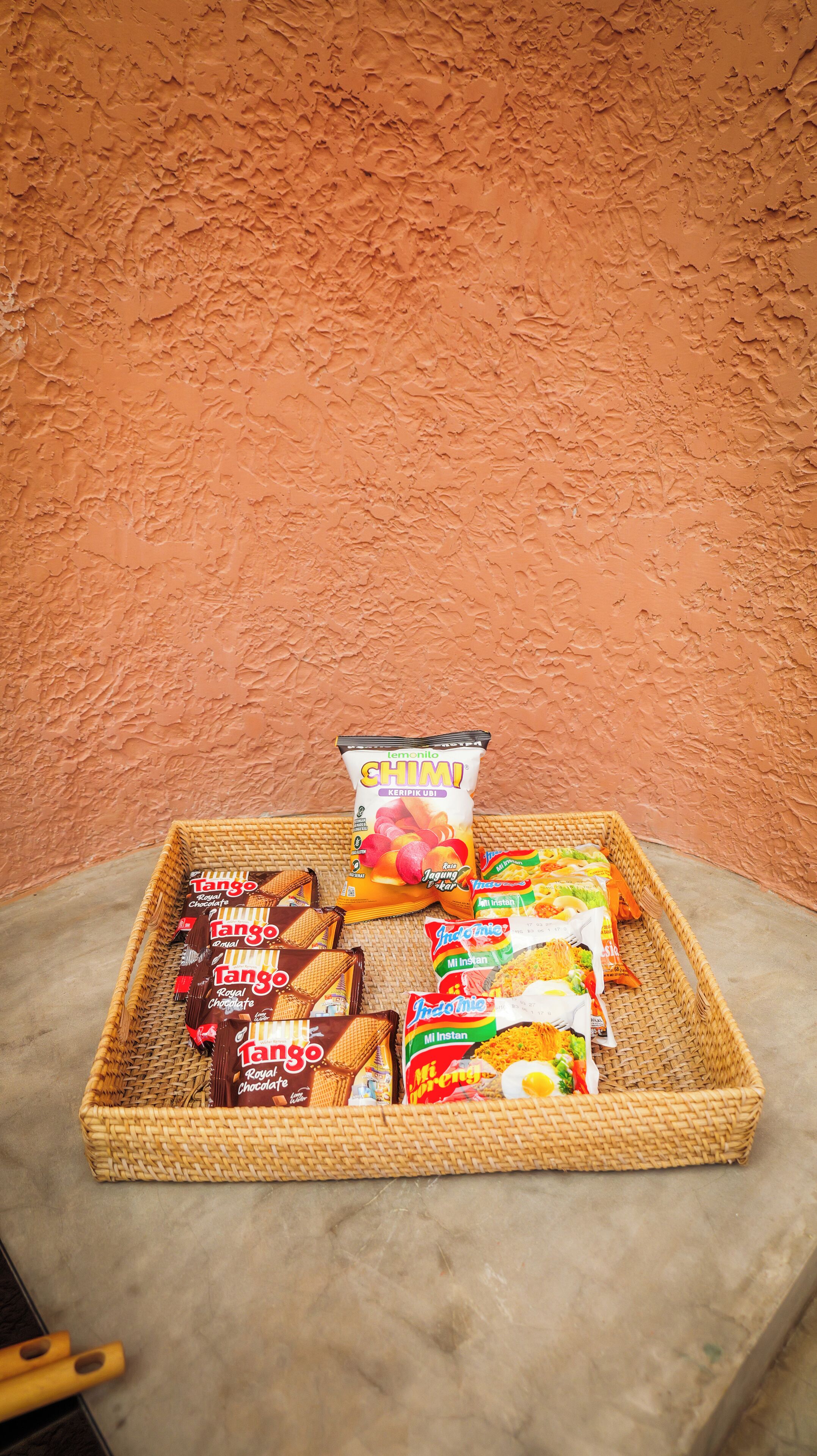A wicker tray with various snacks and a lemon-flavored drink on a concrete surface against a textured wall.