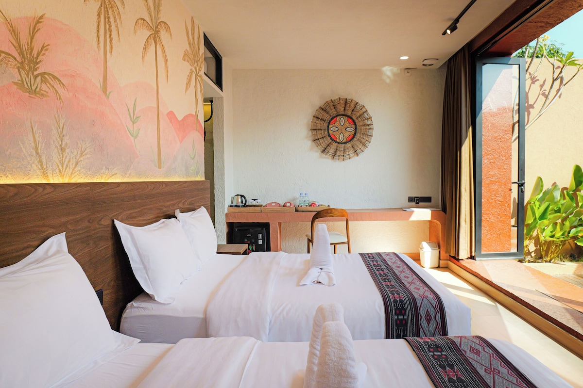 A hotel room with a bed, a desk, a wall-mounted decorative item, and a view of greenery outside.