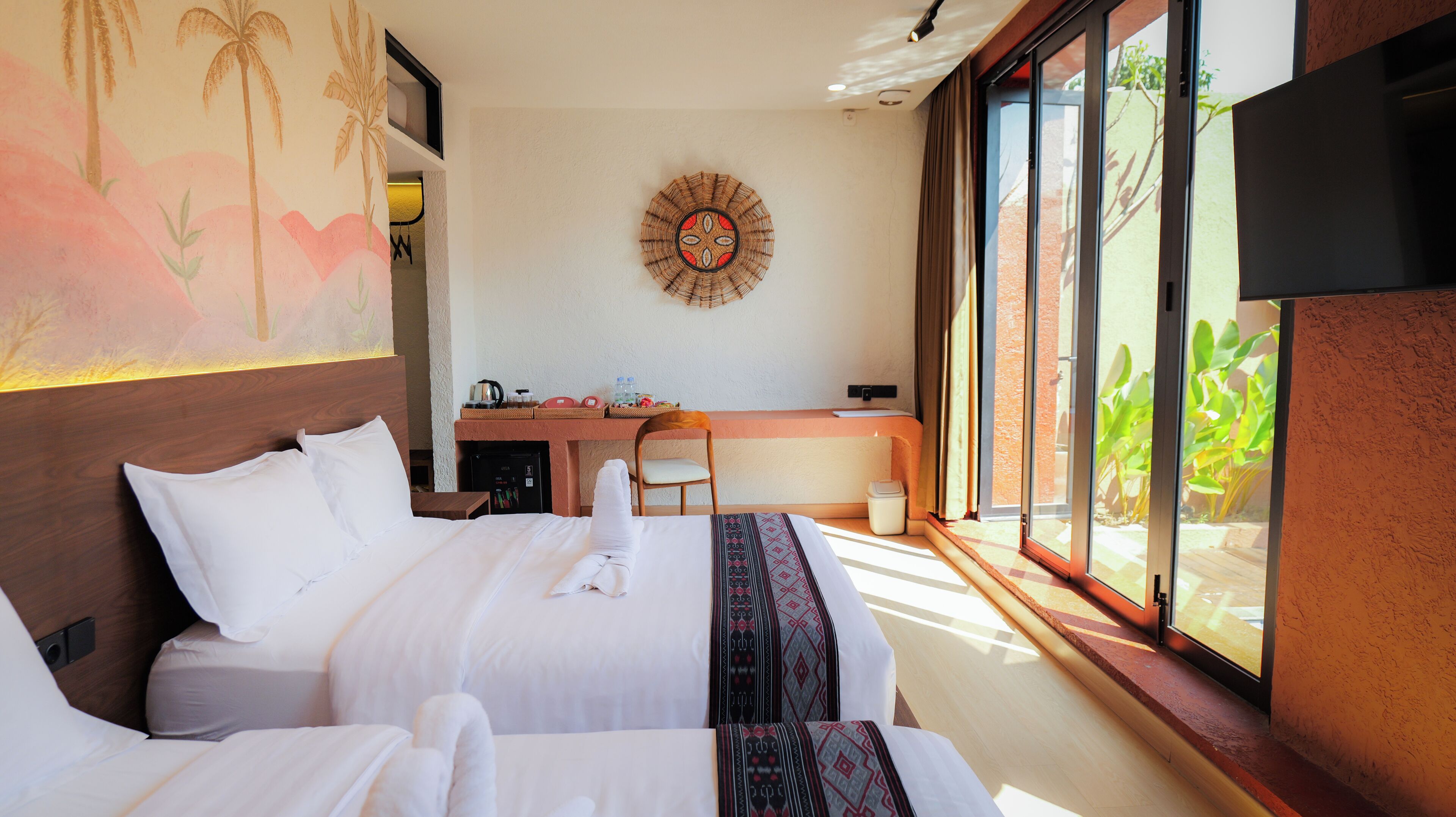 A hotel room with two beds, a desk, a chair, a wall-mounted artwork, and a view of greenery through a window.
