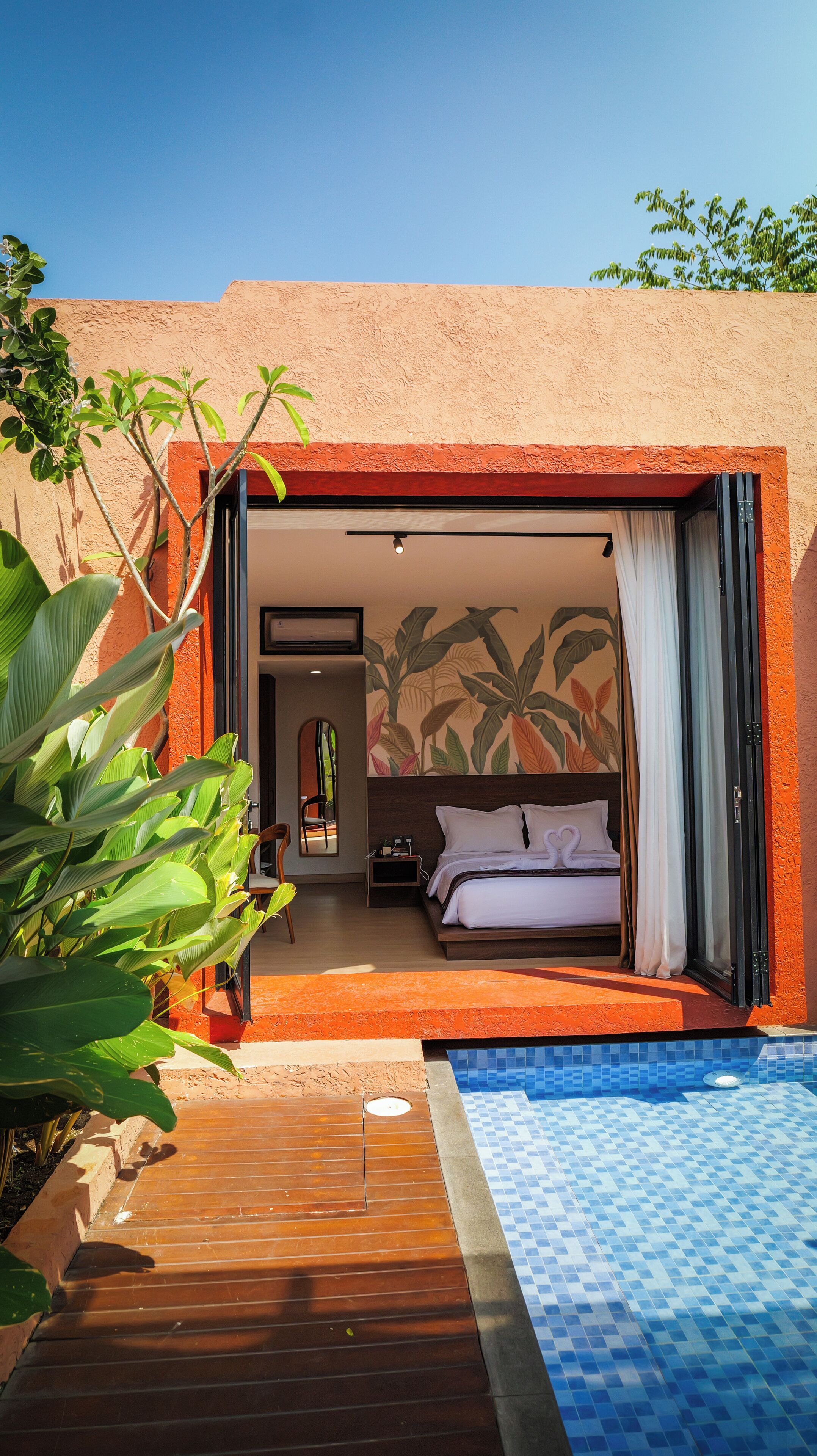 A room with a bed, a small table, and a chair. There is a window with curtains, a wall with a tropical leaf pattern, and a pool with blue tiles.