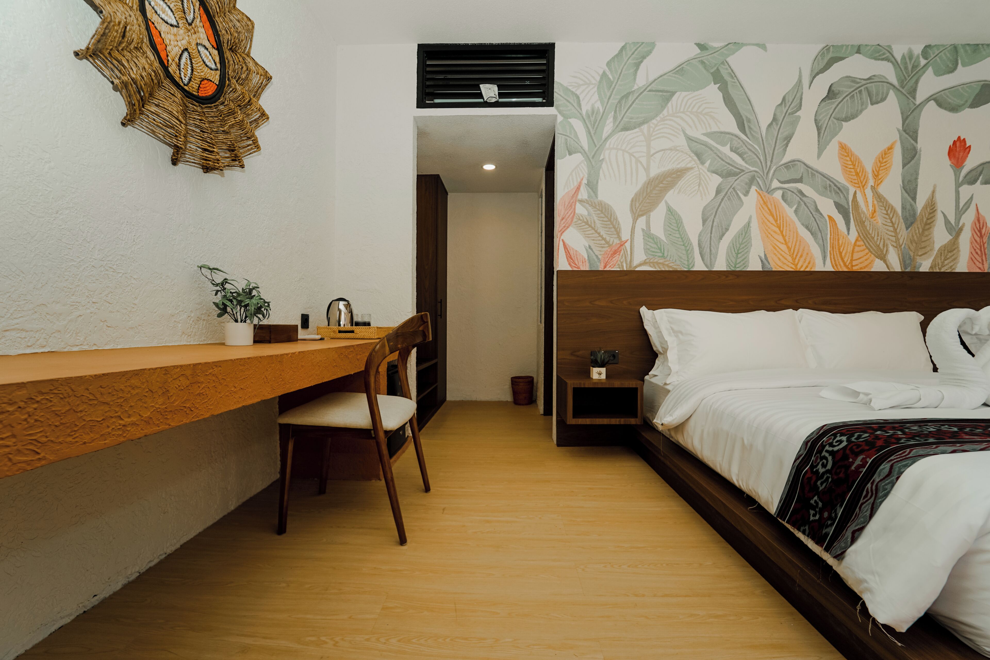 Quarto com escrivaninha de madeira, cadeira, cama com roupa de cama branca e parede com padrão de folha tropical.