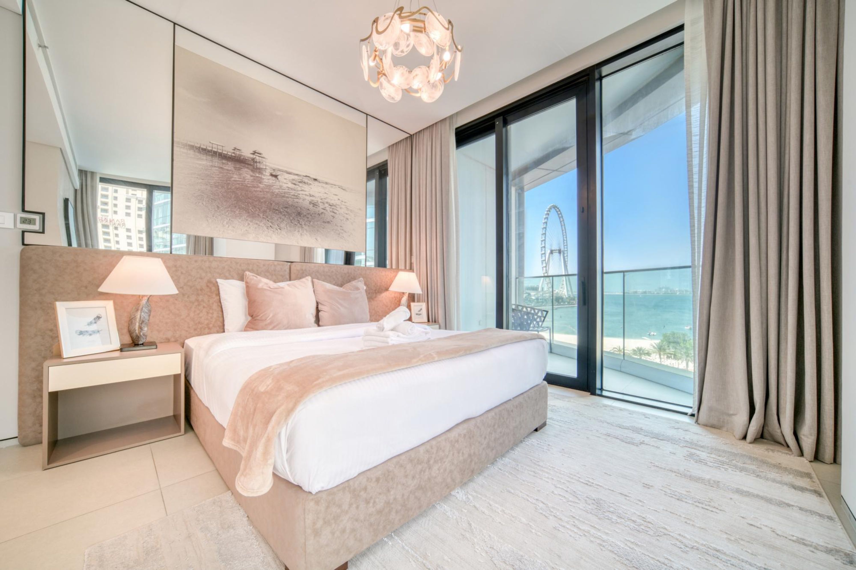 Una habitación de hotel moderna con una cama grande, una mesita de noche, una araña, un ventanal con vistas a una noria y un balcón.