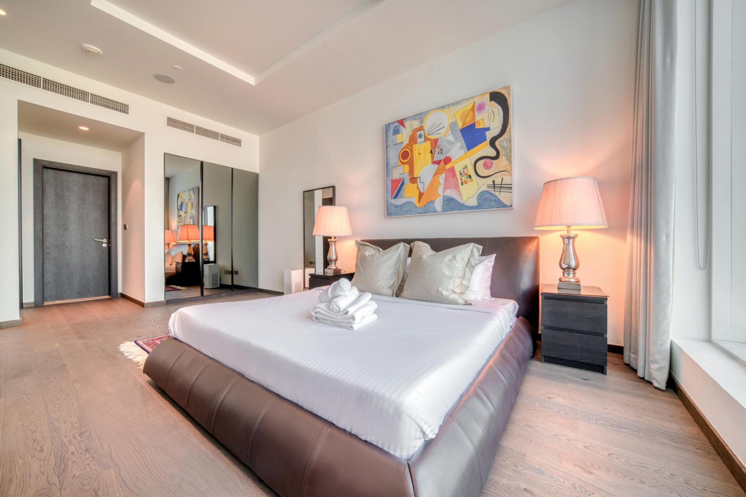 A modern hotel room with a large bed, a painting on the wall, and a mirror.