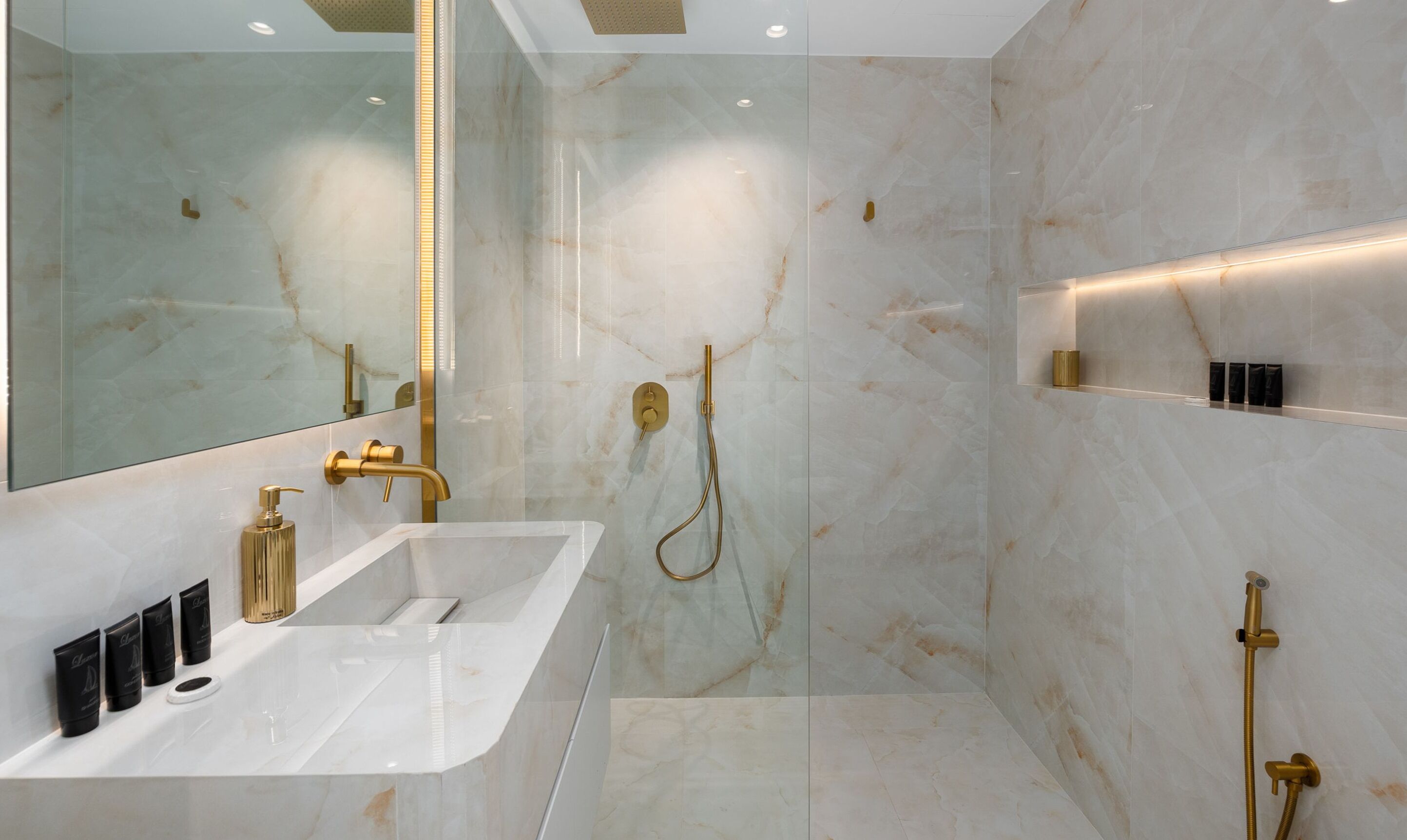 A modern bathroom with a large mirror, a white sink, gold fixtures, and marble walls.