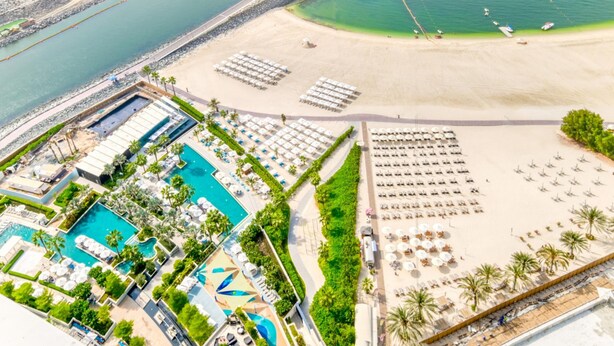 Ein Luftbild zeigt einen Ferienort mit einem großen Pool, Strandbereich und mehreren Liegestühlen.