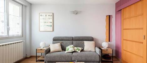 Ein modernes Wohnzimmer mit einem grauen Sofa, einem Holztisch mit Obst, einer Heizung, einem gerahmten Pflanzenmotiv und einer rosafarbenen Wand.