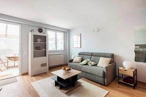 A modern living room with a grey sofa, a coffee table with fruit, a white shelving unit, and a view of the outside. - A39 Exit N*5 Studio: Quiet/Relaxing/Secure (Tréclun)