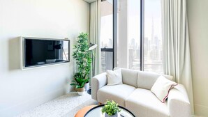 Ein modernes Wohnzimmer mit einer Couch, einem Flachbildfernseher, einer Topfpflanze und Blick auf eine Stadtlandschaft durch große Fenster.
