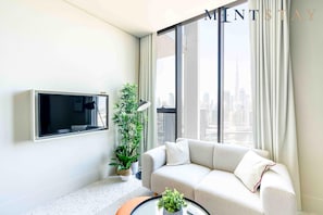 Ein modernes Wohnzimmer mit einer Couch, einem Flachbildfernseher, einer Topfpflanze und Blick auf eine Stadtlandschaft durch große Fenster.
