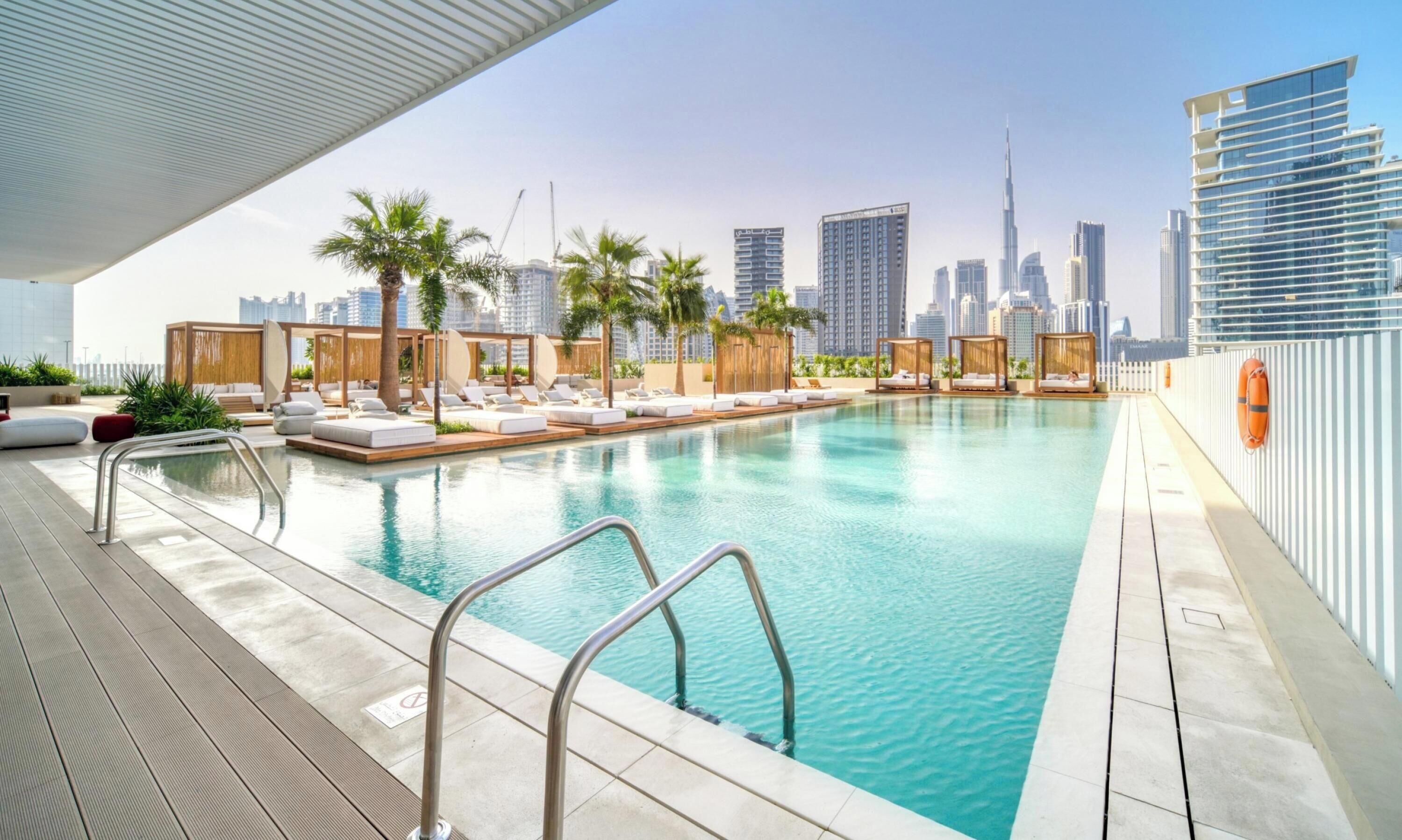 A rooftop pool with lounge chairs and palm trees, overlooking a city skyline with modern buildings.
