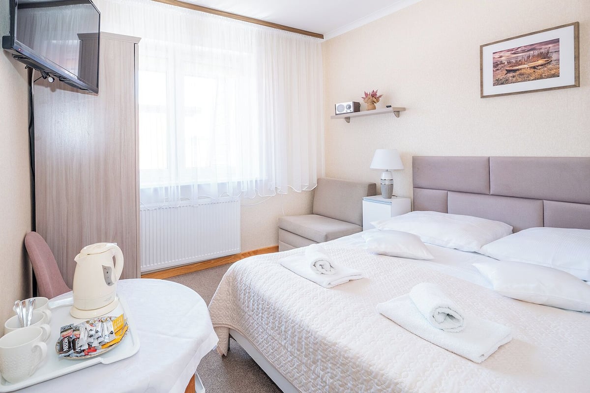 Ein Hotelzimmer mit Bett, Nachttisch mit Lampe, Wandfernseher und Fenster mit leichtem Vorhang.