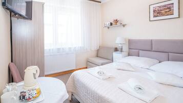 A hotel room with a bed, a bedside table with a lamp, a TV mounted on the wall, and a window with sheer curtains.