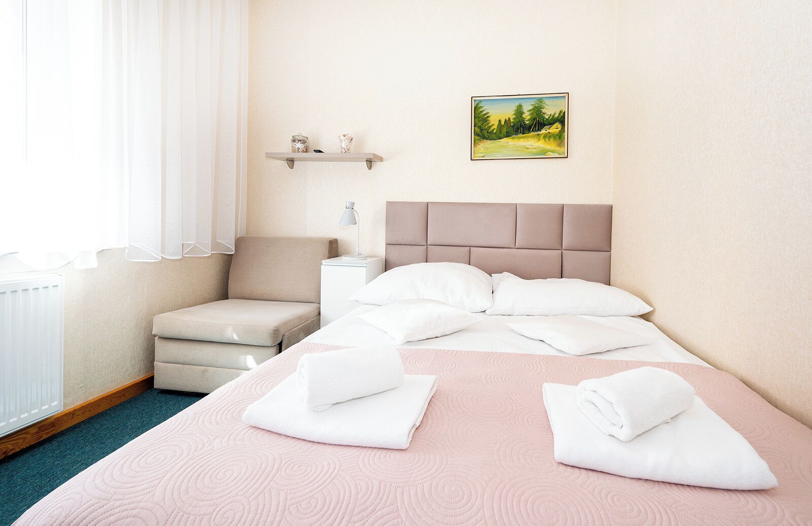 A hotel room with a bed, a chair, a bedside table, a radiator, and a painting on the wall.