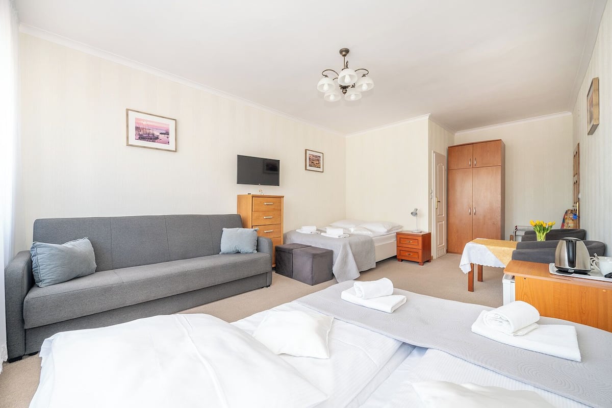 Ein Hotelzimmer mit grauem Sofa, einem Bett mit weißer Bettwäsche, einem hölzernen Schreibtisch mit Wasserkocher und einem an der Wand befestigten Fernseher.