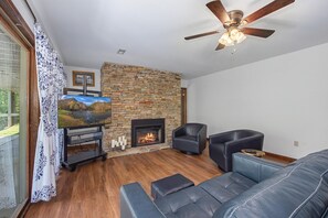 A living room with a stone fireplace, leather sofas, a ceiling fan, and a television on a stand. - River Vibes (Sevierville)