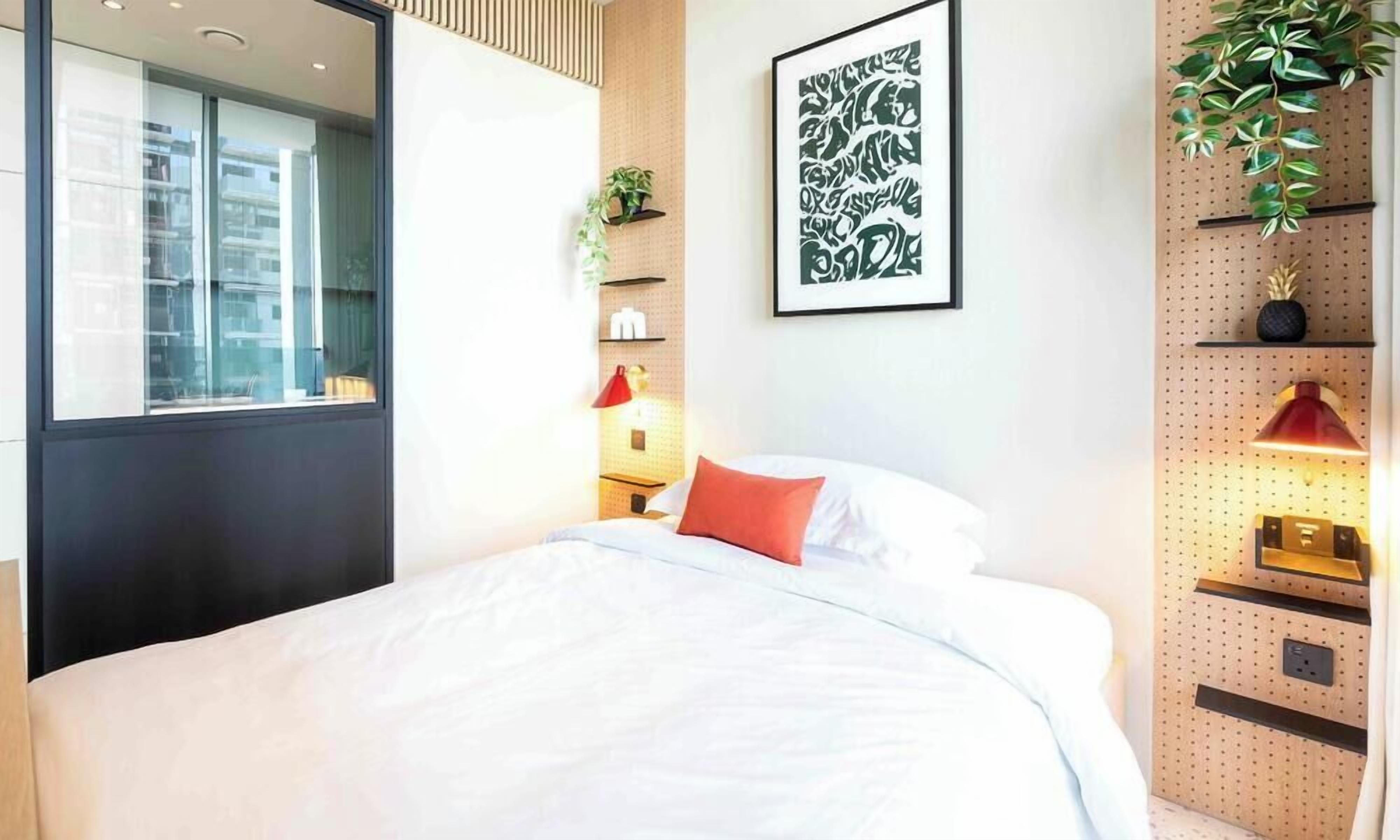 Une chambre d’hôtel moderne avec un grand lit, une fenêtre, une plante fixée au mur, une œuvre d’art encadrée et une étagère murale avec une lampe.