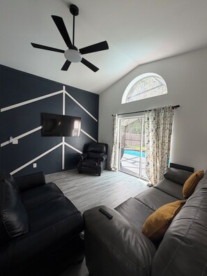 Ein modernes Wohnzimmer mit Deckenventilator, einem flachbildfernseher an der Wand, einem schwarzen Ledersofa und einem großen Fenster mit Vorhängen.