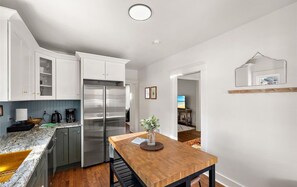 A modern kitchen with white cabinets, a stainless steel refrigerator, a wooden dining table, and a view into the living area.