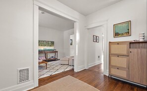 A hallway with a wooden floor, a white wall, a wooden cabinet, and a painting.