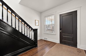 A hallway with a wooden staircase, a dark door, a window with blinds, and a framed picture on the wall.