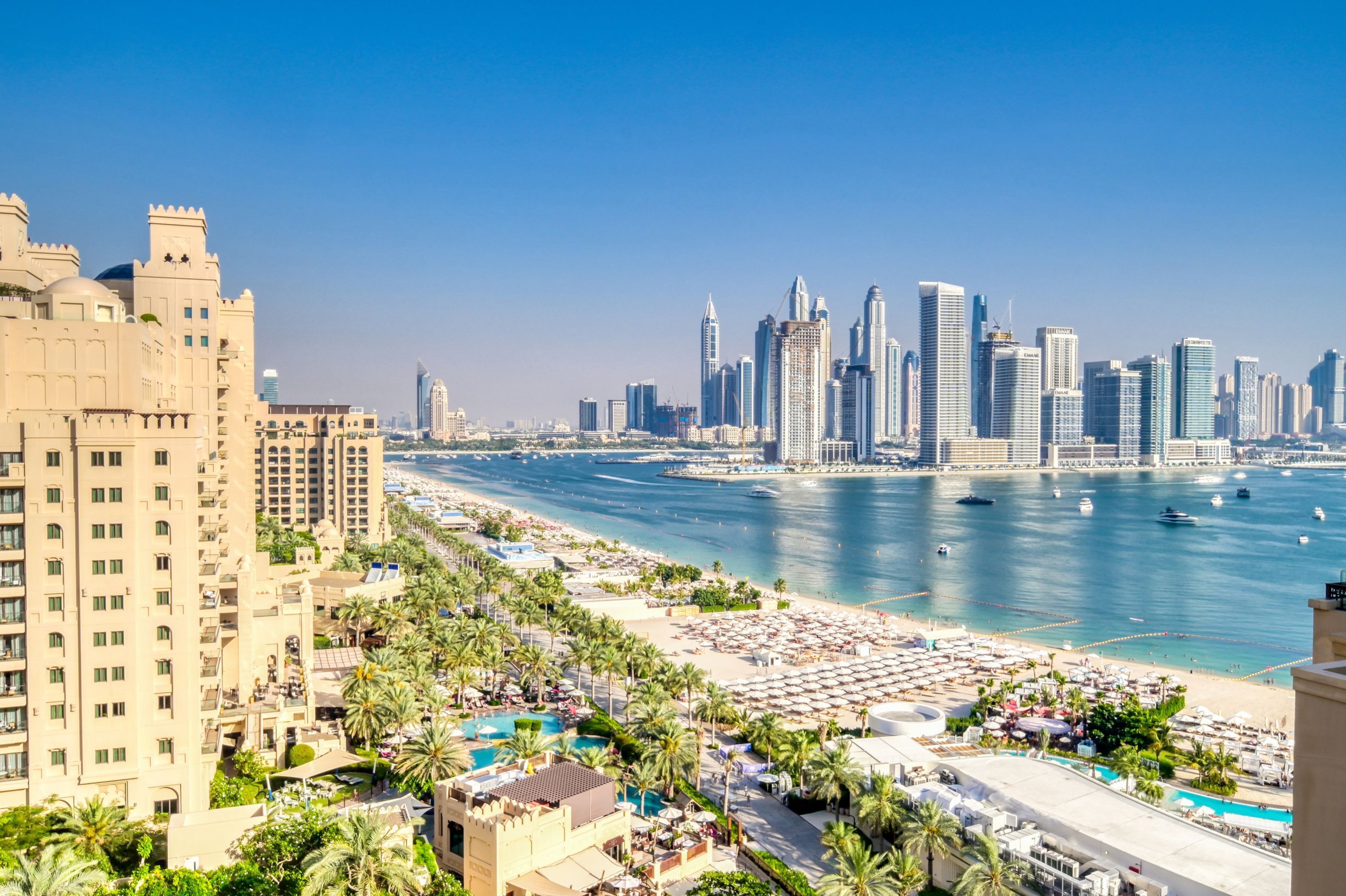 A coastal cityscape with high-rise buildings, a beach with umbrellas, and a clear blue sky.