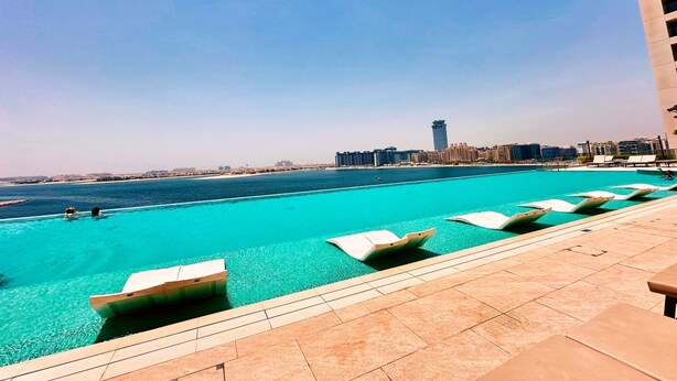 Piscina con sillas de descanso, cielo azul despejado y un paisaje urbano a lo lejos.