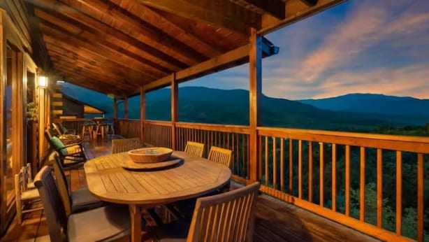 An outdoor wooden deck with a round table and chairs, overlooking a mountainous landscape. - Heaven's Edge (Gatlinburg)