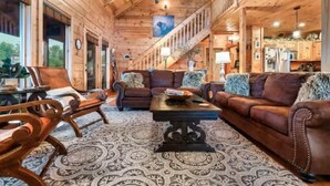 A log cabin interior with a central seating area, wooden walls, and a staircase leading to an upper level. - Heaven's Edge (Gatlinburg)