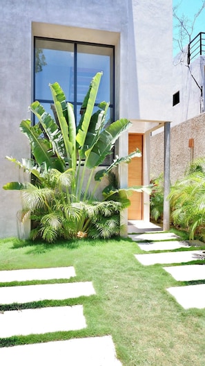 Ein modernes Gebäude mit einem großen Glasfenster, einer Betonfassade und einem gepflegten Garten mit tropischen Pflanzen und einem Kiesweg.