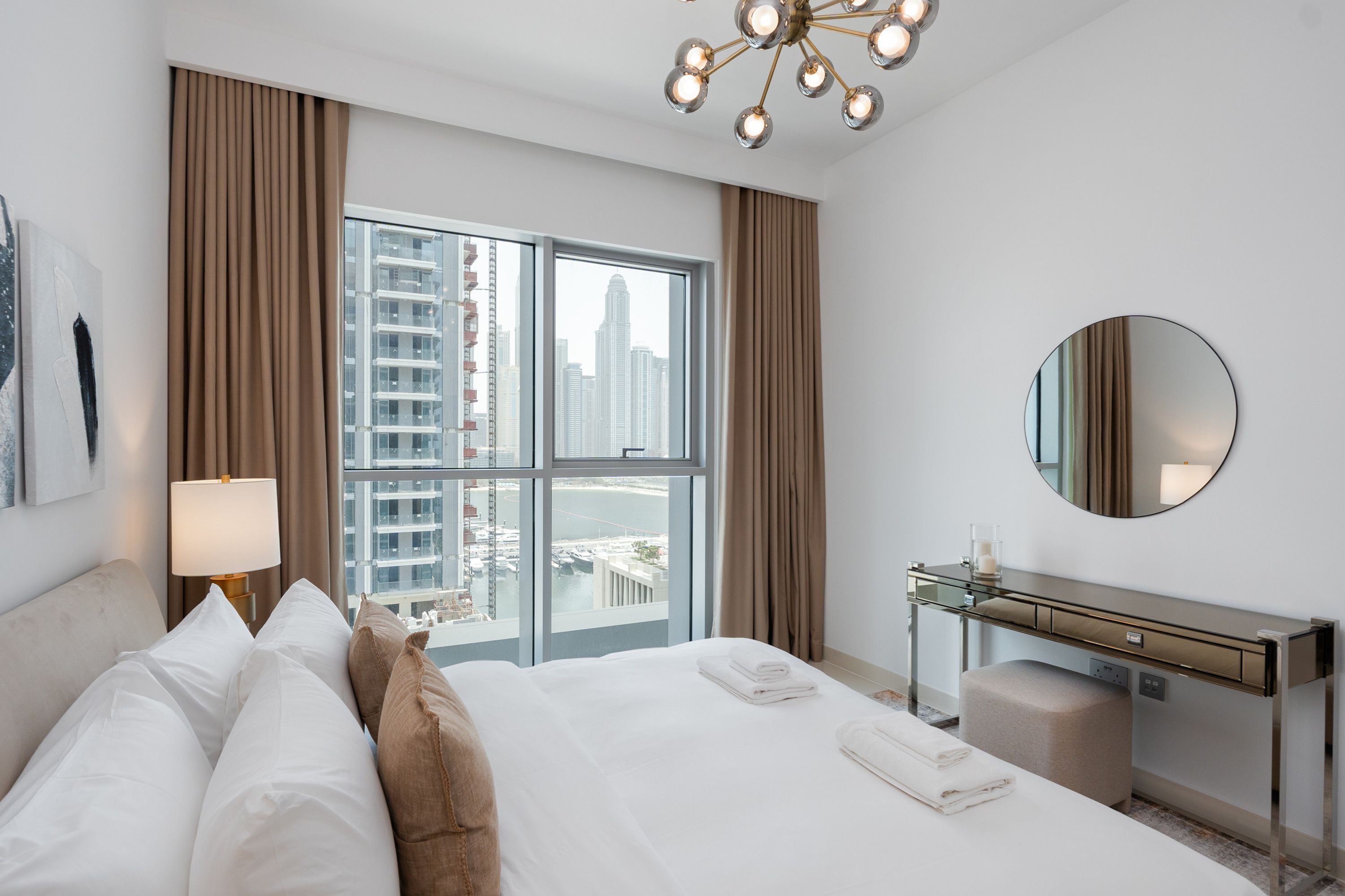 A hotel room with a large bed, a desk, a mirror, and a view of the cityscape through the window.