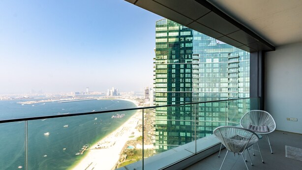 A balcony with a glass railing, two white wire chairs, and a view of a tall building and a beach. - Address JBR Elegant 2BR w/ Beach Access (Dubai)