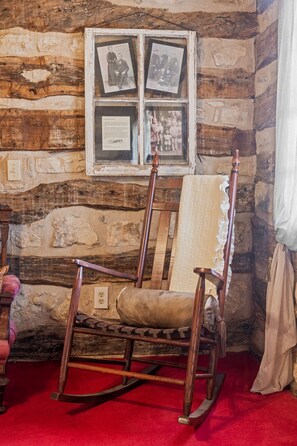 Interior rústico de una cabaña de troncos con una mecedora, un ventanal de madera con fotografías enmarcadas y una alfombra roja.