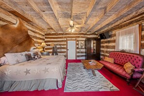 Interior de una cabaña de troncos con una cama, un sofá rojo, una mesa de centro de madera y un ventilador de techo.