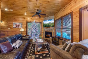 A cozy cabin interior with a leather sofa, a coffee table, a Christmas tree, a fireplace, and a television mounted on the wall. - Eternally Yours (Gatlinburg)
