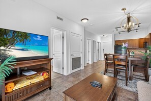 Ein Wohnzimmer mit Fernseher, der eine Strandansicht zeigt, einer Holzunterhaltungsecke, einem Essbereich mit Tisch und Stühlen sowie einer Küche mit Holzausstattung.