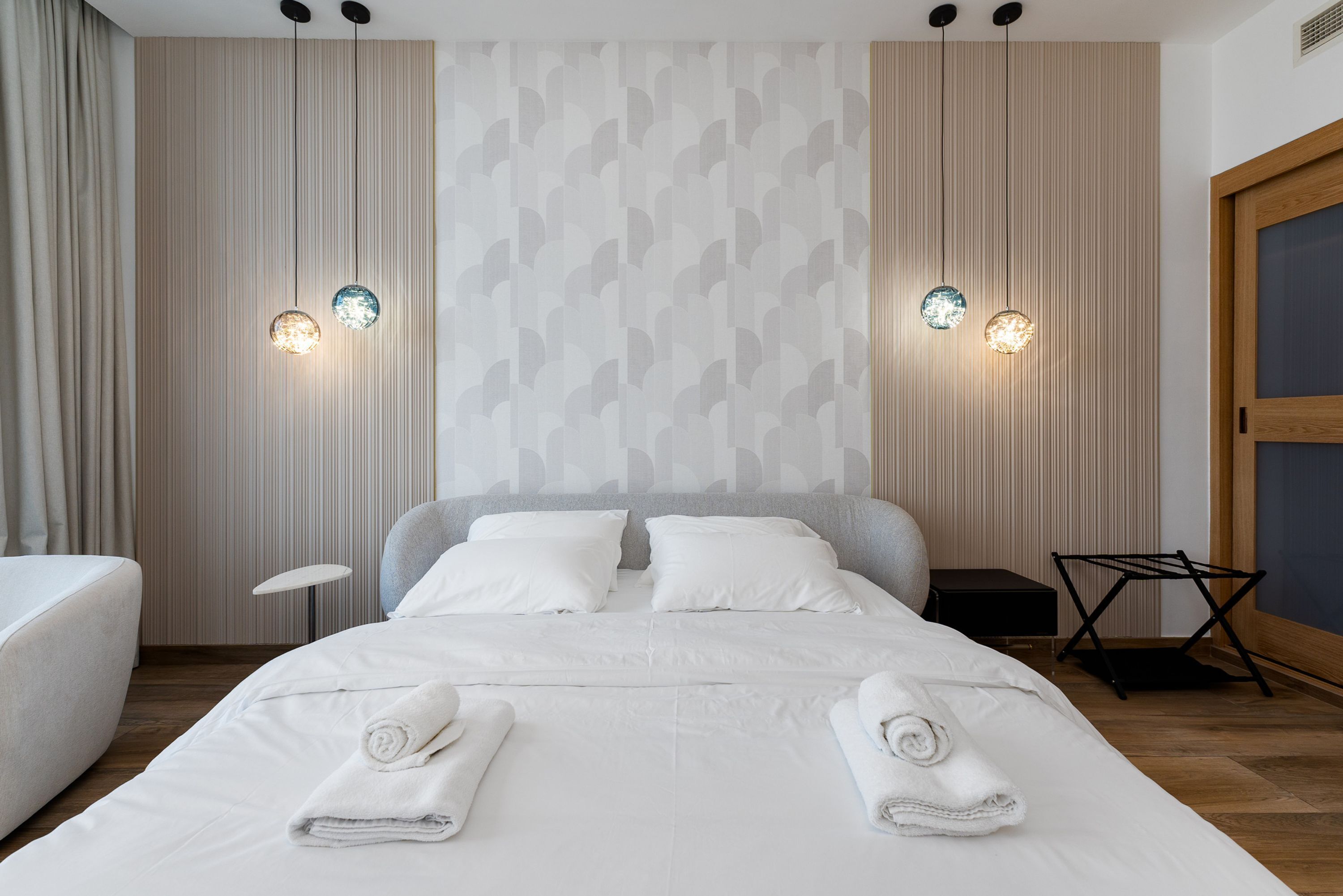 Una cama bien tendida con sábanas y toallas blancas, una mesita de noche y una pared con un diseño geométrico.