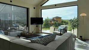 A modern living room with a large sofa, a flat-screen TV, and a dining area with chairs. - New Scandinavian cabin with sea views on Hisøy (Kolbjørnsvik)