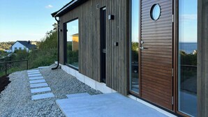 A modern building with a wooden facade, large glass windows, and a gravel pathway leading to the entrance. - New Scandinavian cabin with sea views on Hisøy (Kolbjørnsvik)
