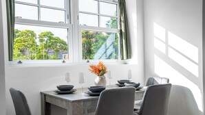 Een eetruimte met een tafel gedekt voor vier personen, een vaas met bloemen en uitzicht op bomen door een raam.