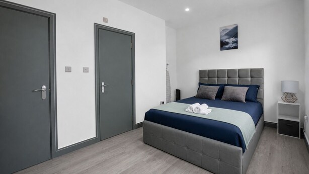 Een slaapkamer met een bed, een grijze hoofdbord, een aan de muur hangend schilderij, en twee deuren.