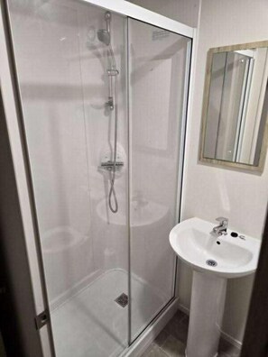 Ducha envuelta en vidrio, con cabezal de ducha de mano y llave montada en la pared. Junto a la ducha, un lavamanos blanco con una sola llave.