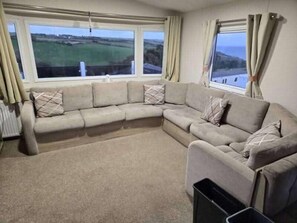 A living room with a beige sectional sofa, two windows with curtains, and a view of a green landscape. - Ty Dee Seaview Escapes (Looe)