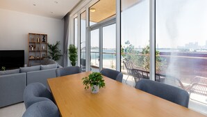 A modern living room with a sofa, a wooden table with a plant, and a TV on a stand. Large windows offer a view of the city and a body of water. - 2BR at the Palm | Balcony, Pool, Gym & Beach (Dubai)