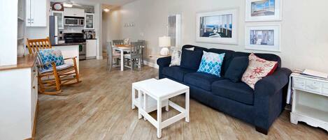 A living room with a dark blue sofa, white coffee table, wooden rocking chair, and a dining area with a table and chairs.