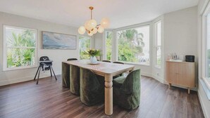 A dining area with a wooden table, green upholstered chairs, a hanging light fixture, and large windows with a view of greenery. - Sandcastle | Steps to Beach | Pool | Golf Cart (Anna Maria)