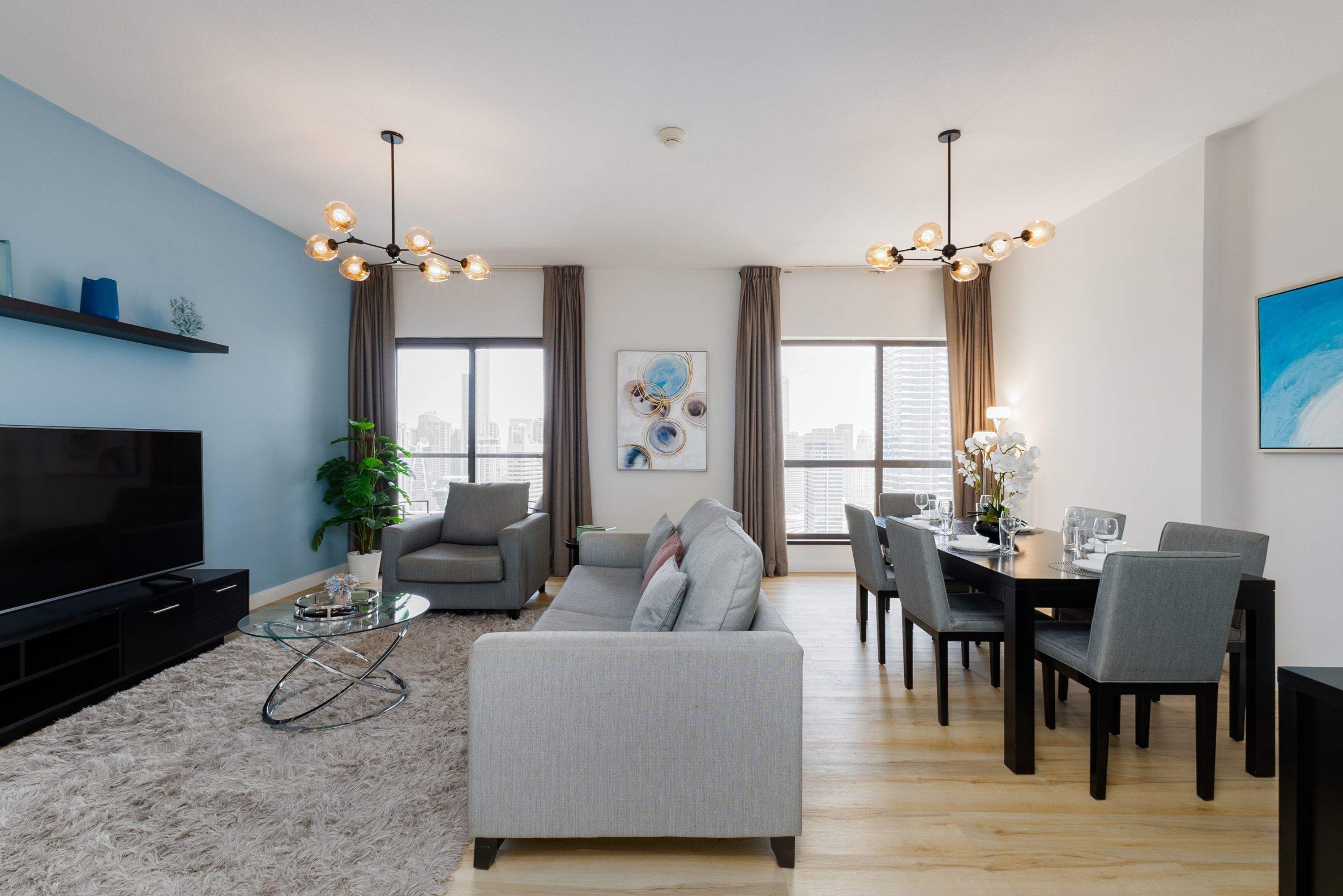 Ein modernes Wohnzimmer mit Flachbildfernseher, grauem Sofa, Esstisch für sechs Personen und Stadtblick durch große Fenster.