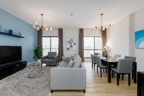 Ein modernes Wohnzimmer mit Flachbildfernseher, grauem Sofa, Esstisch f ür sechs Personen und Stadtblick durch große Fenster.