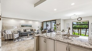 Uma cozinha moderna com bancadas de granito, uma sala de jantar com mesa e cadeiras brancas, e uma sala de estar com sofá e poltronas.